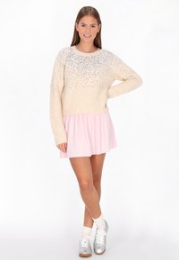 Pulover bej cu embellismente sclipitoare la decolteu, asortat cu o fustă plisată roz deschis. Modelul poartă adidași argintii și șosete albe.