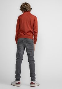 Maglione rosso lavorato a maglia con colletto alto, abbinato a jeans slim-fit grigi. Sneakers bianche con dettagli rossi. Design semplice e pulito.