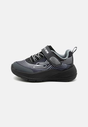 Zapatos deportivos en negro y gris con parte superior de malla, líneas blancas curvas y una correa de velcro sobre los cordones. Suela de goma texturizada.