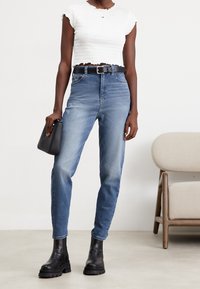 Frau trägt ein weißes kurz geschnittenes Oberteil, hochgeschnittene blaue Jeans mit schwarzem Gürtel, schwarze Knöchelstiefel und hält eine schwarze Handtasche.