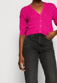 Cárdigan de punto rosa brillante con cuello en V, mangas cortas y patrón de diamantes; presenta grandes botones transparentes y dobladillo acanalado. Se lleva con jeans negros.