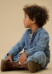 Gaiši zils džinsa kombinezons ar garām piedurknēm un pogu aizdari, valkāts kopā ar brūnām potīšu zābakiem, sēžot uz bēša fona.