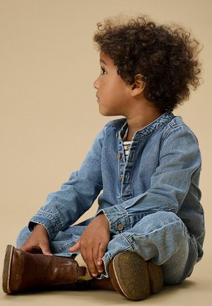 Tuta in denim azzurro chiaro con maniche lunghe, abbottonatura sul davanti, indossata con stivaletti marroni, seduta su uno sfondo beige.