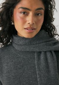 Temno sivek pleteni pulover z visokim, zloženim ovratnikom. Mehak teksturiran material z sproščenim krojem. Model ima kodraste lase in nevtralen naravni videz.