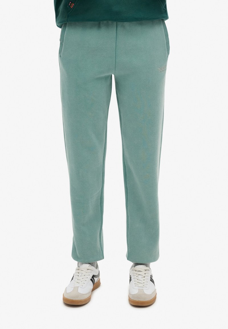 Pantalones de chándal suaves de color verde menta con corte recto, cintura elástica, bolsillos laterales y una marca sutil en el muslo izquierdo. Combinados con zapatillas blancas.