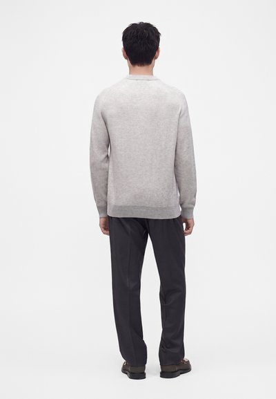 Homme debout de dos, portant un pull gris clair, un pantalon noir et des chaussures foncées sur un fond blanc uni.