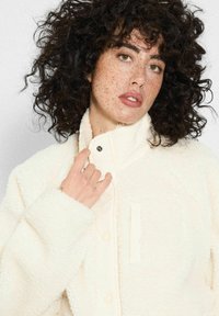 Kobieta z kręconymi ciemnymi włosami i piegami, ubrana w kremową polarow�ą kurtkę, trzymającą kołnierz jedną ręką na jednolitym tle.