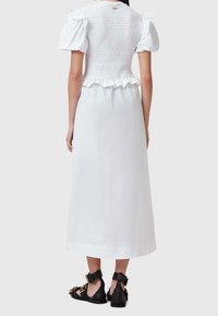 Vestido maxi blanco con un corpiño fruncido, mangas cortas abullonadas y una cintura con volantes. Combinado con zapatos negros con detalles dorados.