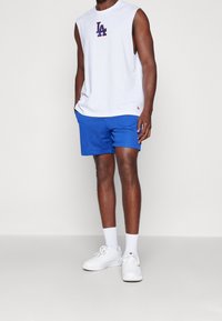 Bílá bezrukávová košile s červeným logem "LA", doplněná modrými šortkami z pružné síťoviny a bílými teniskami s bílými kotníkovými ponožkami. Neformální sportovní outfit.