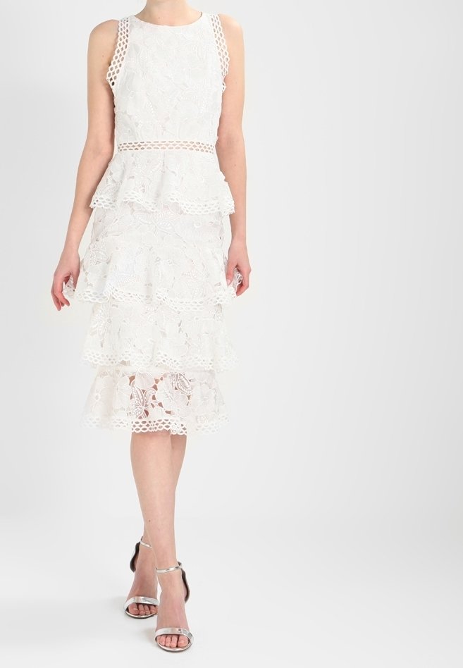 Femme portant une robe blanche sans manches en dentelle avec des volants à étages et des talons ouverts argentés, debout devant un fond clair uni.