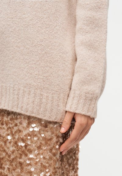 Ανοιχτό μπεζ πουλόβερ με απαλό, υφασμάτινο φινίρισμα και ριμπ στην άκρη, σε συνδυασμό με μια λαμπερή φούστα με παγέτες σε ζεστή καφέ απόχρωση.