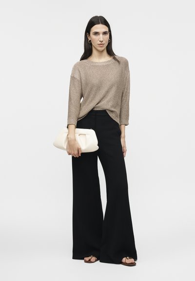 Femme aux longs cheveux foncés porte un pull en maille beige, un pantalon noir à jambes larges, des sandales et tient une pochette blanche sur un fond uni.