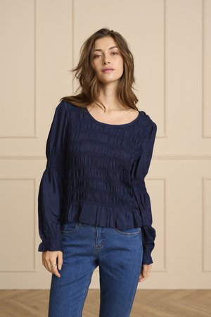 Mladá žena s vlnitými hnedými vlasmi má na sebe námornícky modrú nariasenú blúzku s dlhými rukávmi a modré džínsy, stojí v béžovej miestnosti s panelmi.