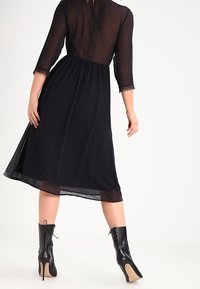Schwarzes halbtransparentes Kleid mit Dreiviertelärmeln, Punktmuster, taillierter Taille und fließendem Rock, kombiniert mit schwarzen High-Heel-Stiefeln.