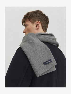 Grey knitted scarf draped over the shoulder, featuring a textured pattern and a navy label with white text. Worn with a dark coat.