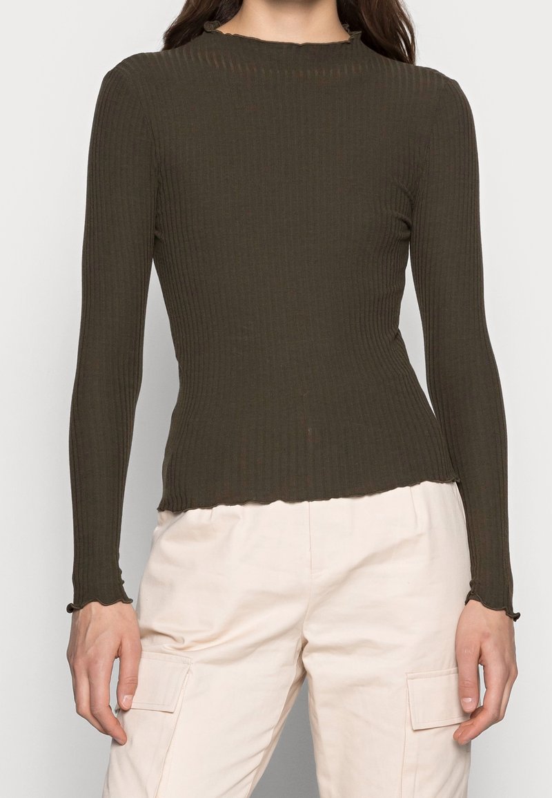 Femme portant un haut moulant à manches longues, côtelé, de couleur olive foncé, associé à un pantalon beige clair avec des poches cargo latérales.