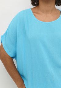 Personne aux cheveux bouclés portant un haut bleu clair à manches courtes et coupe ample, avec un col rond, debout devant un fond blanc uni.