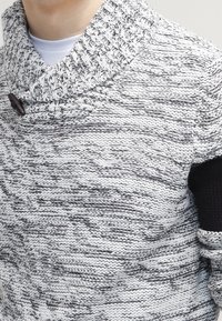 Stickad tröja i grå och vit melerad mönster med krage, utrustad med en träknapp och svarta accenter på ärmarna. Texturerad tyg.