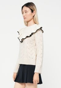 Cream knitted sweater with a textured chevron pattern and a large ruffled collar edged in black. Paired with a black pleated skirt.