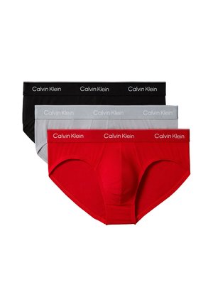 Hombre de pie descalzo frente a un fondo liso, lleva calzoncillos claros Calvin Klein y la mano izquierda tocando la parte posterior de su cuello.