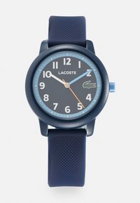 Reloj con correa de silicona azul marino, esfera redonda azul marino, marcadores de hora en blanco y azul, logo de Lacoste y segundero naranja. Superficie de la correa texturizada.