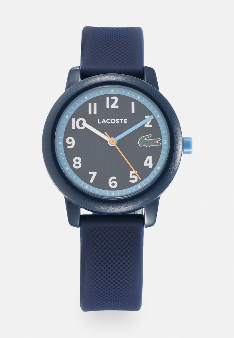 Reloj con correa de silicona azul marino, esfera redonda azul marino, marcadores de hora en blanco y azul, logo de Lacoste y segundero naranja. Superficie de la correa texturizada.