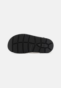 Svart gummisåle med et teksturert mønster av spor og rygger for grep. Glatte kanter og en fremtredende logo.