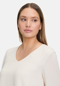 Blusa a scollo a V di un leggero beige, realizzata in un tessuto morbido. Maniche lunghe con una texture soffice, vestibilità elegante, senza motivi o accenti visibili.