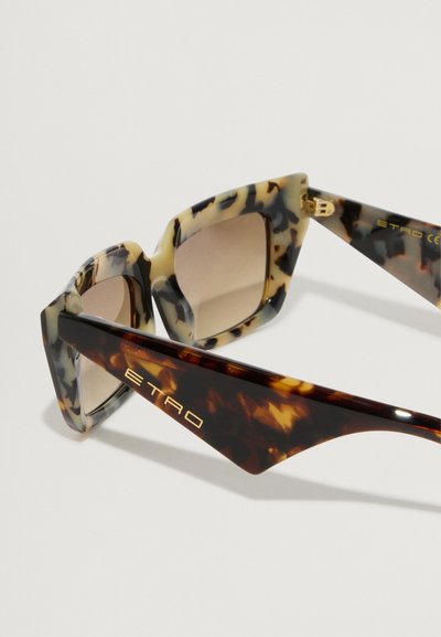 Óculos de sol com um design geométrico e oversized. A armação apresenta um padrão tartaruga em bege e castanho. As lentes são ligeiramente tonalizadas. Marca em ouro.