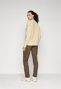 Woman with dark curly ponytail wearing beige knitted sweater, brown cargo pants, and cream sneakers, standing and looking back over her shoulder.