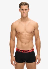 Schwarze Baumwollboxer mit einem roten elastischen Bund, der mit "SUPERDRY" Branding versehen ist. Das Design ist enganliegend und hat eine glatte Textur.