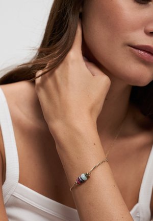 Woman wearing a gold chain bracelet with blue, purple, and pink beads, touching her neck, wearing a white sleeveless top.