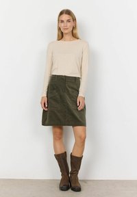 Beige pikaaedega topp, mille juurde on sobitatud tume roheline puuvillane seelik, mis sisaldab esikülje taskuid, ja pruuni kõrge säärega saapad.