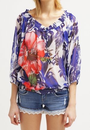 Mujer que lleva blusa floral transparente con grandes flores rojas y hojas azules, combinada con pantalones cortos de mezclilla con bordes de encaje.