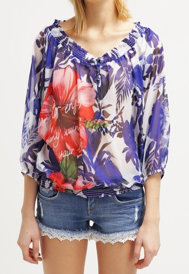 Femme portant un chemisier floral transparent avec de grandes fleurs rouges et des motifs de feuilles bleues, associé à un short en jean avec bordure en dentelle.