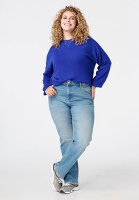 Blauwe gebreide trui, lichtgewassen spijkerbroek, grijze sneakers. Model heeft krullend haar en staat met één voet over de andere gekruist, glimlachend.