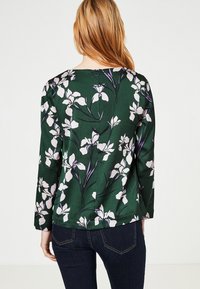 Mörkgrön blus med vitt och lila blommönster, långa ärmar, rund hals, slät textur och veckdetalj på ryggen.