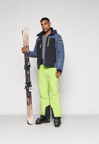 Veste de ski avec des panneaux bleus et noirs, accents jaune fluo ; associée à un pantalon de ski vert vif. Skis en main, chaussures de ski noires visibles.