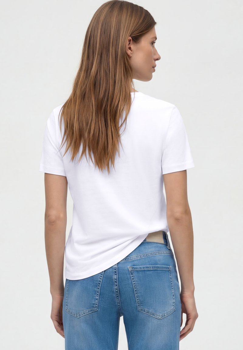 Femme aux cheveux longs bruns portant un T-shirt blanc à manches courtes uni et un jean bleu, tournée de dos contre un fond neutre.