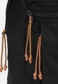 Zaino in tessuto nero con cordini marroni e punte in plastica, dotato di una tasca con zip e superficie liscia.