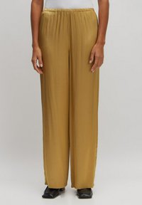 Weite Hosen aus einem glatten, goldbraunen Stoff mit einem elastischen Bund, kombiniert mit schwarzen, eckigen Schuhen.