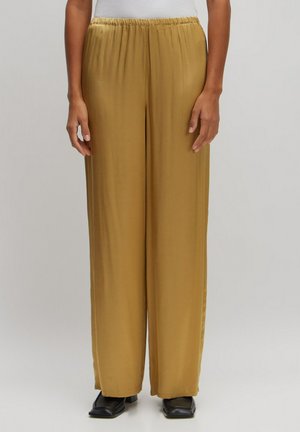 Wijdere broek in een zachte, goudbruine stof met een elastische tailleband, gecombineerd met zwarte schoenen met een vierkante neus.