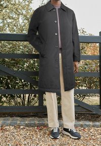 Long manteau noir avec fermeture à boutons, porté sur un pull violet clair, associé à un pantalon crème et des chaussures de sport noires. Fond de feuillage vert.