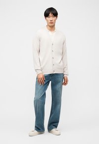 Νεαρός άνδρας, φορώντας μπεζ ζακέτα με κουμπιά, λευκό πουκάμισο, μπλε τζιν παντελόνι με φαρδιά μπατζάκια και λευκά αθλητικά παπούτσια, στέκεται μπροστά σε απλό λευκό φόντο.