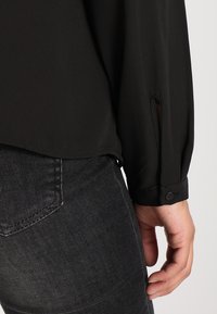Blouse noire en tissu léger et lisse, à manches longues avec des détails de poignet boutonné et une coupe décontractée, associée à un jean sombre.
