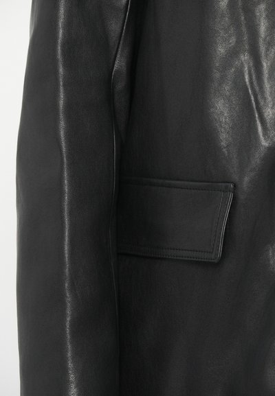 Κοντινή άποψη μανικιού μαύρης δερμάτινης ζακέτας και πλάγιας τσέπης με ορατές ραφές και λεπτή υφή σε λείο υλικό.