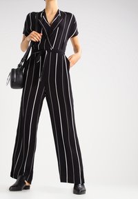 Černobílý pruhovaný overal s krátkými rukávy, zavázáním v pase a širokými nohavicemi, doplněný malou kulatou černou taškou a černými nazouvacími botami.