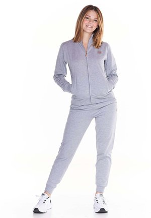 Une femme souriante porte un survêtement gris clair zippé, avec un pantalon assorti et des baskets blanches, sur fond blanc.