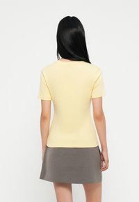 Kortermet, gul topp med en tettsittende design, laget av mykt materiale, kombinert med et grått skjørt, sett fra bak.