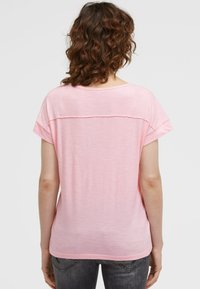 Woman with curly brown hair wearing a light pink short-sleeve shirt and dark gray jeans, shown from the back against a plain background.